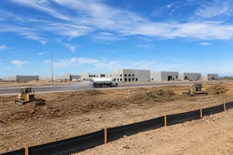 Two machines working windrows of mulched brush on a multi-acre commercial clearing site with silt fence, a water truck, and tilt-up warehouse shells rising across the road