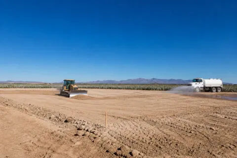 Small dozer leveling tan fill over a staked desert homesite while a water truck dampens the dust along the pad edge