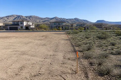 Half-cleared building lot on a subdivision edge with smooth bare dirt on one side, native creosote on the other, and survey stakes with orange flagging