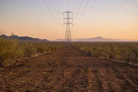 Long mulched utility easement running straight across creosote flats with steel transmission towers receding toward the horizon in evening light