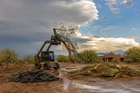 Grapple loader lifting a wind-snapped tree on desert acreage after a monsoon storm with debris piles raked and puddles still standing